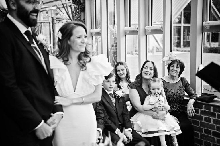 B&W documentary photo of front row of wedding guests looking towards bride and groom during the wedding at Syrencot.