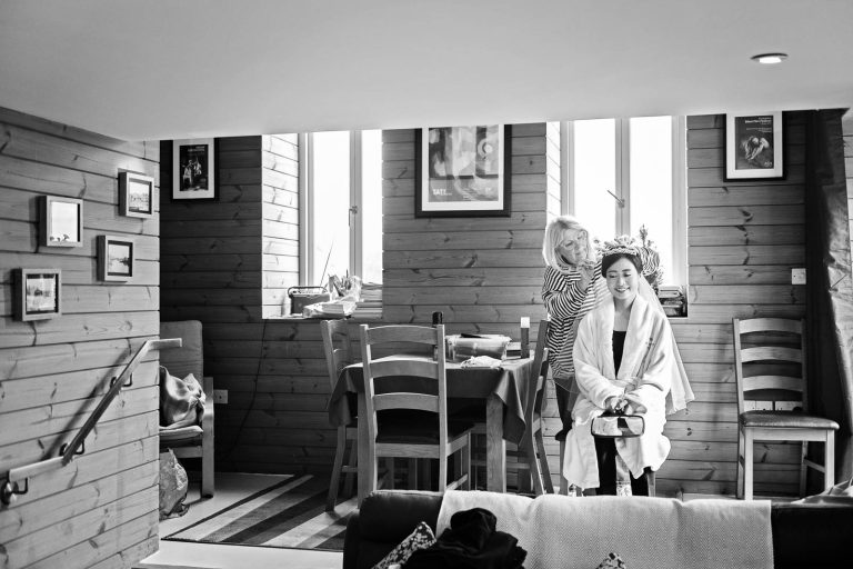 Bride having her hair done in a wooden slatted living room. Using natural light. Bride is smiling. Photograph by Blooming Photography.