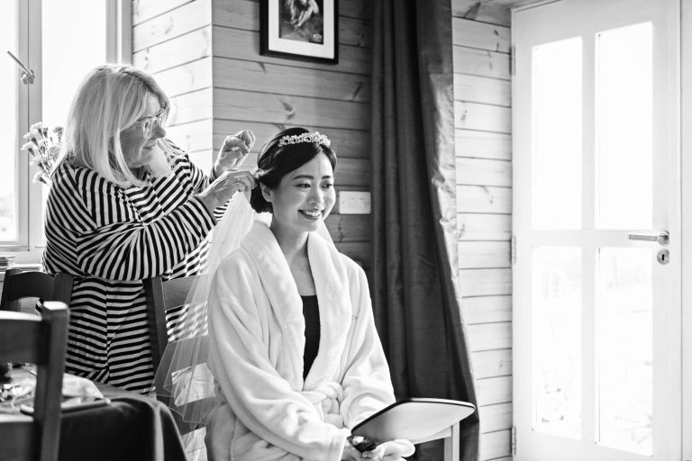 Bride having her wedding veil added in a wooden slatted living room. Using natural light. Bride is smiling. Photograph by Blooming Photography.