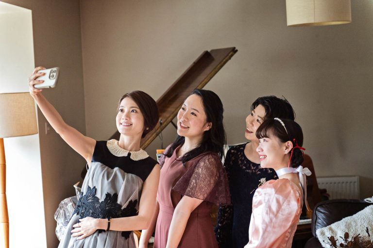 A group of girls smile at a phone as they take a selfie together. Candid photograph by Blooming Photography.