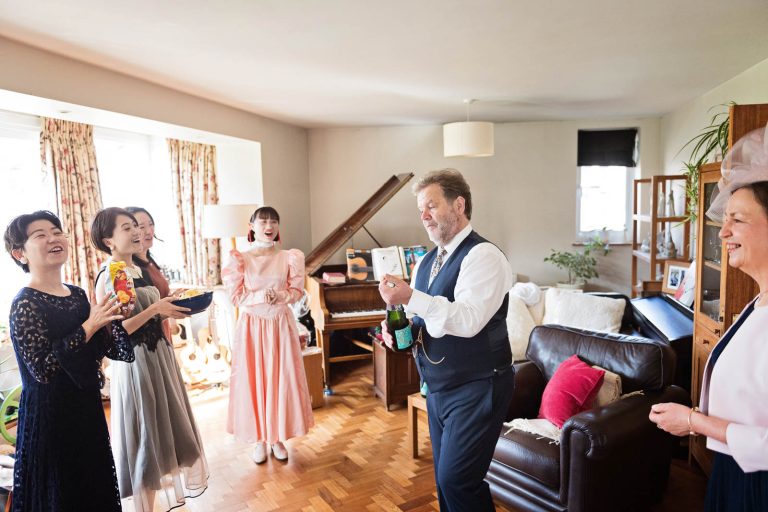 Father of the groom opens up a bottle of fizz as mum and bridesmaids cheer. In a living room. Candid photograph by Blooming Photography.