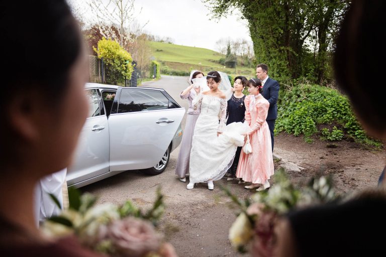 Candid photo of bride getting out of wedding car being helped by her bridesmaids. Candid photograph by Blooming Photography.