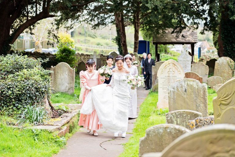 Bride walks down church path. Candid photograph by Blooming Photography.