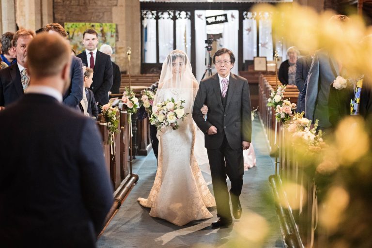 The bride walking down the wedding isle of St George Parish Church, Easton-In-Gordano. The groom and all the guests watch. Candid photograph by Blooming Photography.