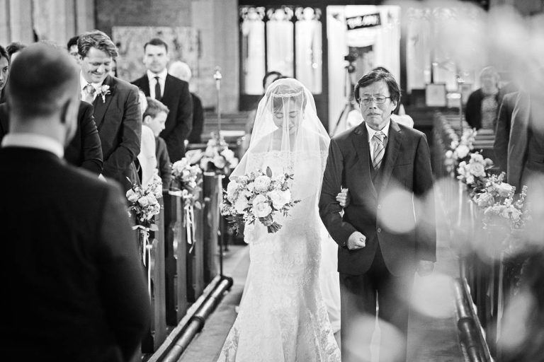 The bride walking down the wedding isle of St George Parish Church, Easton-In-Gordano. The groom and all the guests watch. Black and white candid photograph by Blooming Photography.