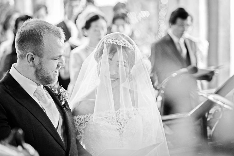 The bride and groom sneak a glance at each other during a wedding hymn. Black and white candid photograph by Blooming Photography.
