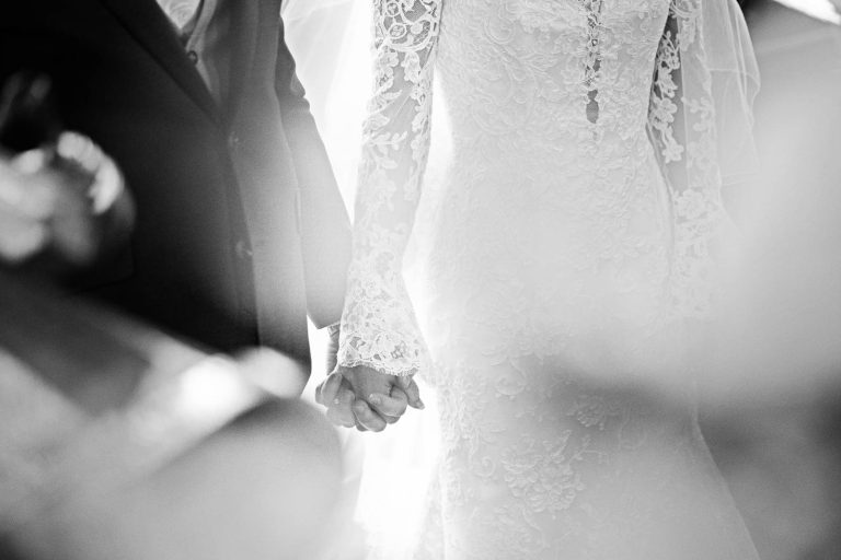 The bride and groom hold hands during a wedding ceremony. Black and white candid photograph by Blooming Photography.