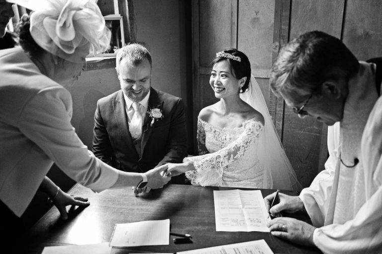 Mother of the bride looks at the bride and her ring. Bride is smiling, vicar is signing the wedding certificate. Groom is smiling. Story telling photograph by Blooming Photography.