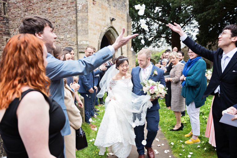 Bride and groom walk through their guests arch whilst being showered with confetti. Story telling photograph by Blooming Photography.