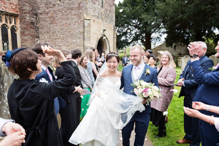 Candid photograph of bride and groom exiting the church whilst being showered with confetti. Guests smiling. Story telling photograph by Blooming Photography.