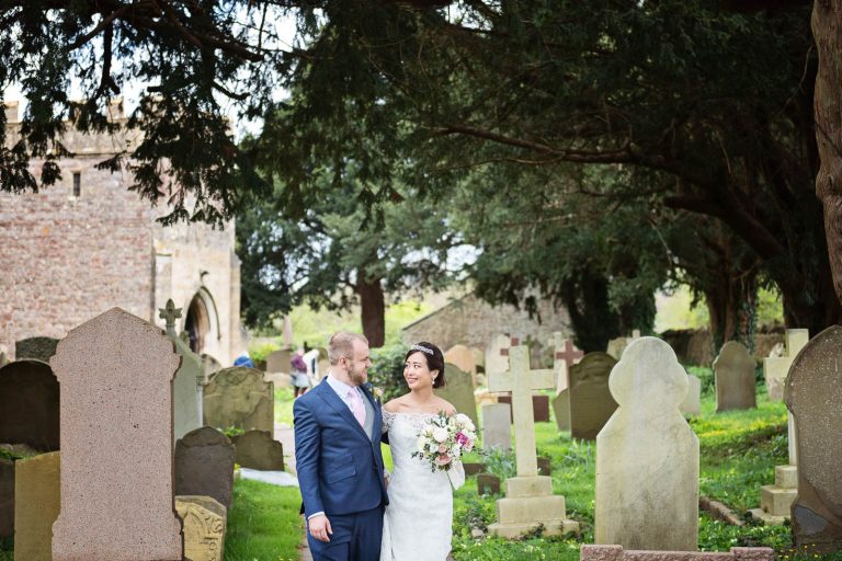 A bride and groom stand in the grounds with graves either side of them. Candid photograph by Blooming Photography.