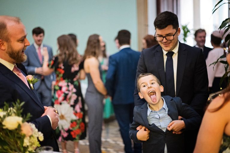 Candid wedding photo of young boy sticking his long tongue out. The boy is being held by a guest for balance. Candid photograph by Blooming Photography.