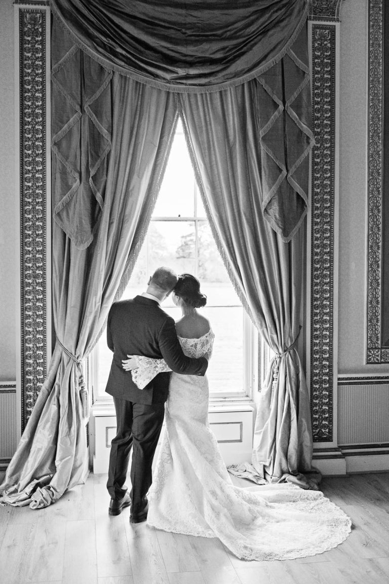Portrait photo of Bride and groom standing together in the window of the tapestry room in Leigh Court. Candid black and white photograph by Blooming Photography.