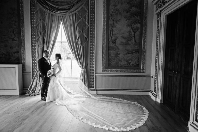 Bride and groom standing together in the window of the tapestry room in Leigh Court. The brides large veil is laid out. Candid black and white photograph by Blooming Photography.