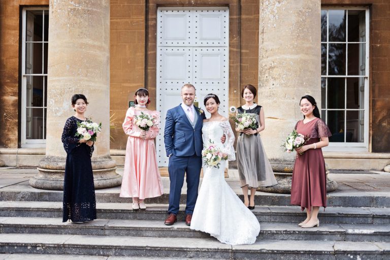 Formal photograph of bride and groom and bridesmaids stand on the steps of Leigh Court, Bristol, holding their wedding flowers. Candid photograph by Blooming Photography.