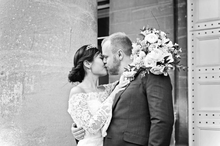 Classic photo of bride and groom kissing next to the pillar at Leigh Court. The bride is holding her wedding flowers over the grooms shoulder. Candid photograph by Blooming Photography.