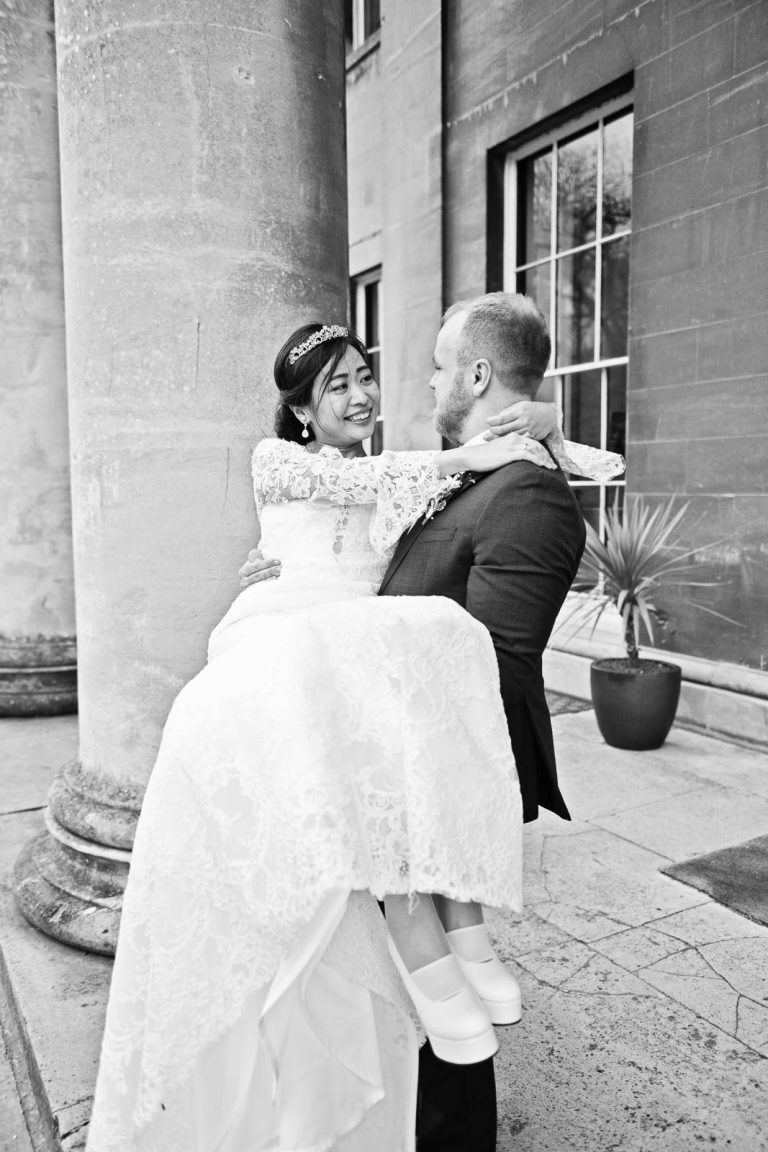 Portrait photo of the Groom lifting his bride up into his arms in the middle of the stone pillars at Leigh Court, Bristol. Black and white candid photograph by Blooming Photography.