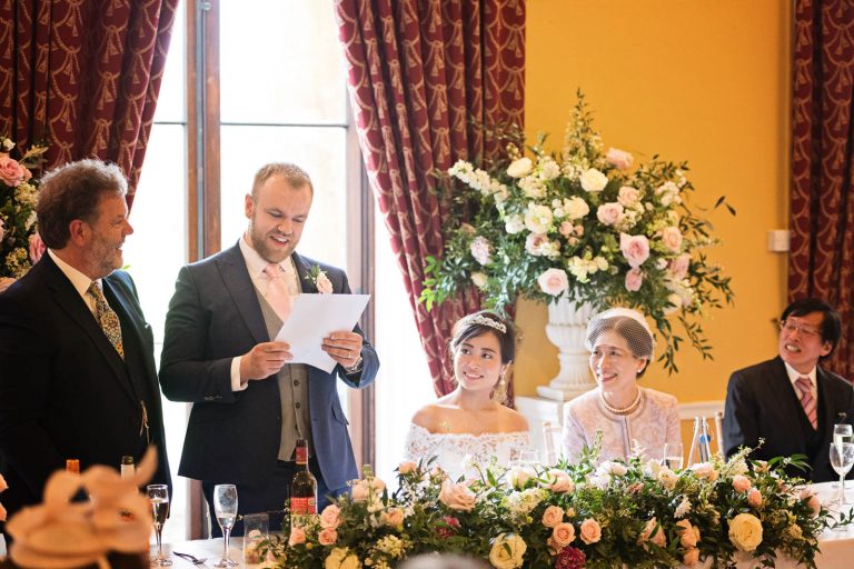 Groom translates the father of the grooms speech into Japanese. Top table look on smiling. In the library in Leigh Court. Photo by Blooming Photography.