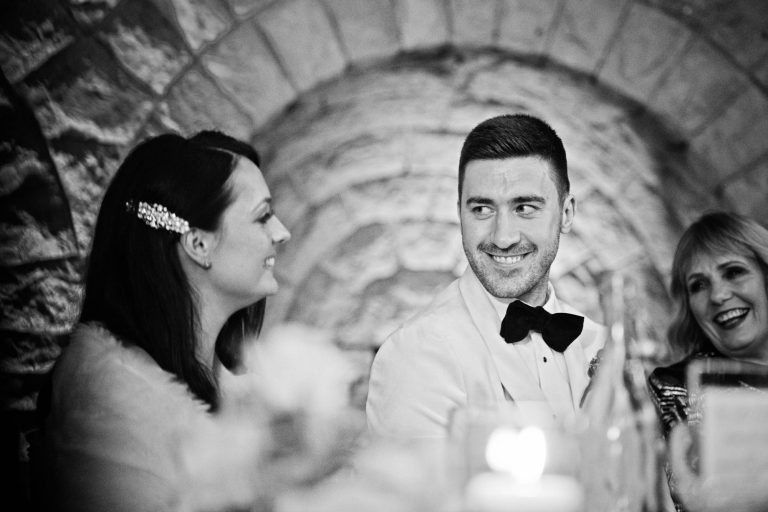 Candid b&w photo during wedding speeches of bride and groom smiling at each other.