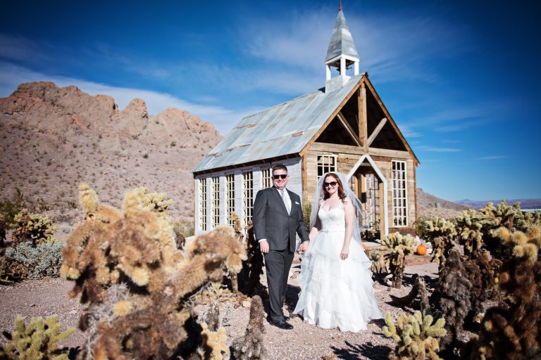 Bride and groom stand amongst cacti and in front of a chapel in the Nevada desert at Nelson Ghost Town.