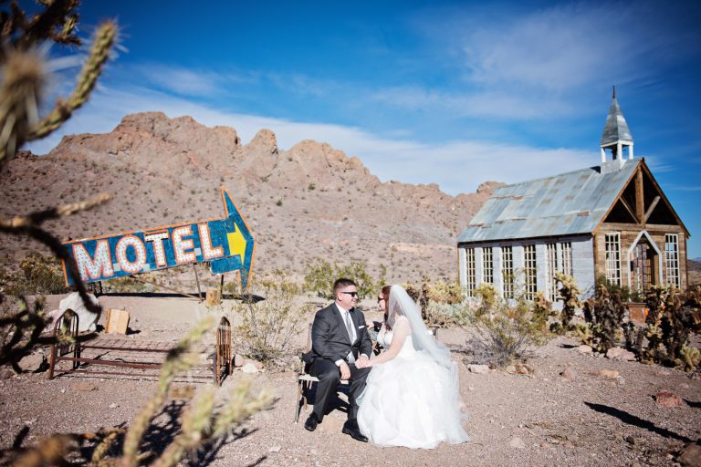 Bride and groom sit amongst cacti and in front of a chapel in the Nevada desert at Nelson Ghost Town. A Motel Arrow sign points towards the bride and groom and the chapel.