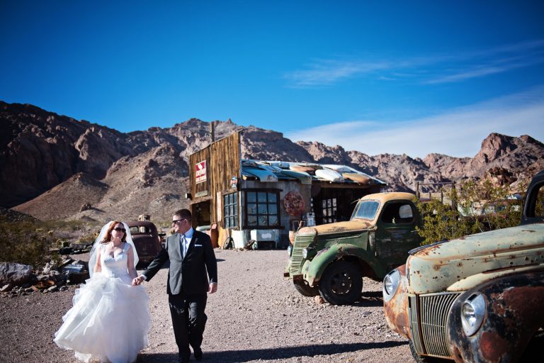 Bride and groom walk and in hand away from an old gas station with 1950's old trucks and cars. Photograph by Blooming Photography, photographed at Nelson Ghost Town.