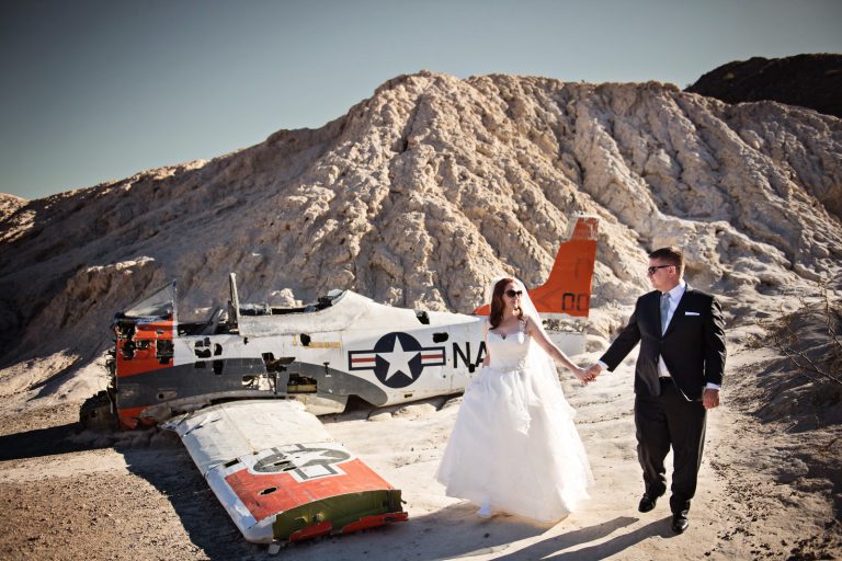 Bride and groom walk and hold hands next to an old US Navy Fighter Plane. Photograph by Blooming Photography, photographed at Nelson Ghost Town.