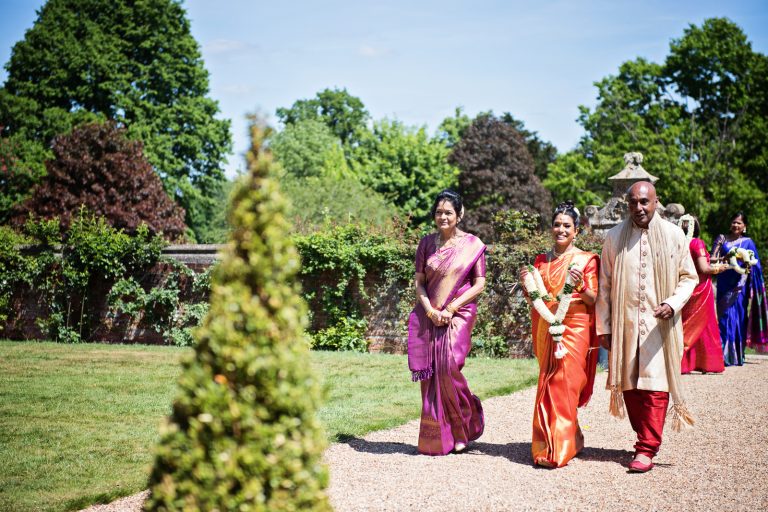 Bride and her mother and father walk together, bride is holding a garland during Hindu ceremony at North Mymms Park, candidly photographed by Blooming Photography.