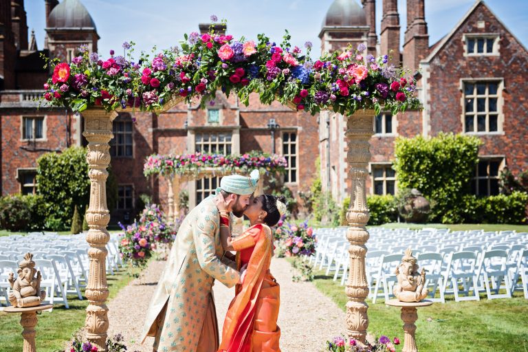 Bride and groom kiss under arch after Hindu ceremony at North Mymms Park, candidly photographed by Blooming Photography.