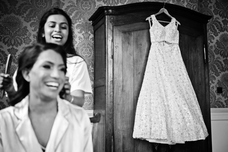 Bride and her hairdresser laugh out of focus with a wedding dress hung up on the wardrobe in focus, black and white photo taken at North Mymms Park, candidly photographed by Blooming Photography.