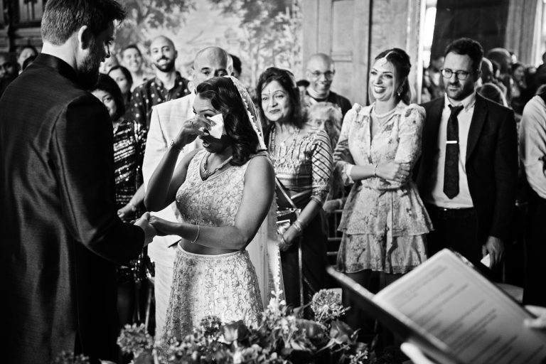 A black and white photo of a bride using a tissue to wipe the tears away from her eyes as she stands at the alter saying her wedding vows. Her family are smiling behind her. Photographed at North Mymms Park, by Blooming Photography.