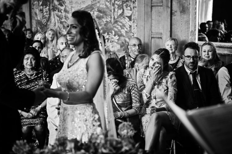 Sister of the bride wipes her eye from tears as she looks towards her sister, who is getting married (& smiling). Black and white image. Photographed at North Mymms Park, by Blooming Photography.