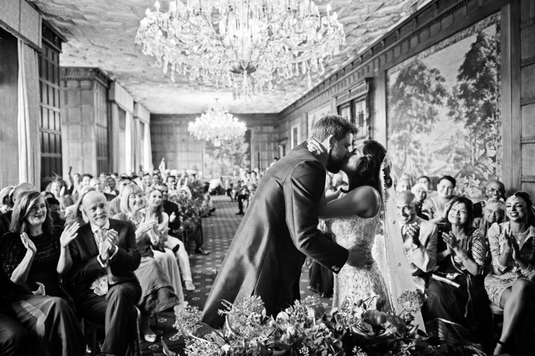 Photograph of bride and groom kissing during the wedding ceremony. Wedding guests in the backgroud clapping and smiling. Black and white image. Photographed at North Mymms Park, by Blooming Photography.)