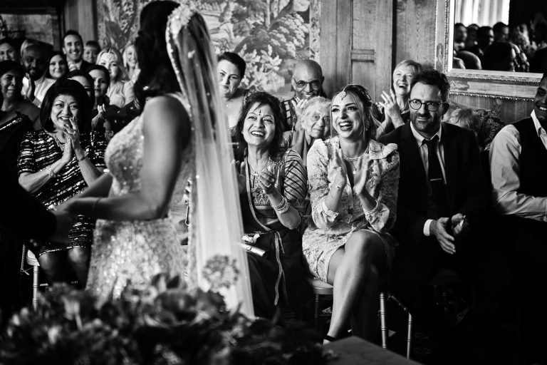 Wedding guests in the backgroud clapping and smiling. Black and white image. Photographed at North Mymms Park, by Blooming Photography.)
