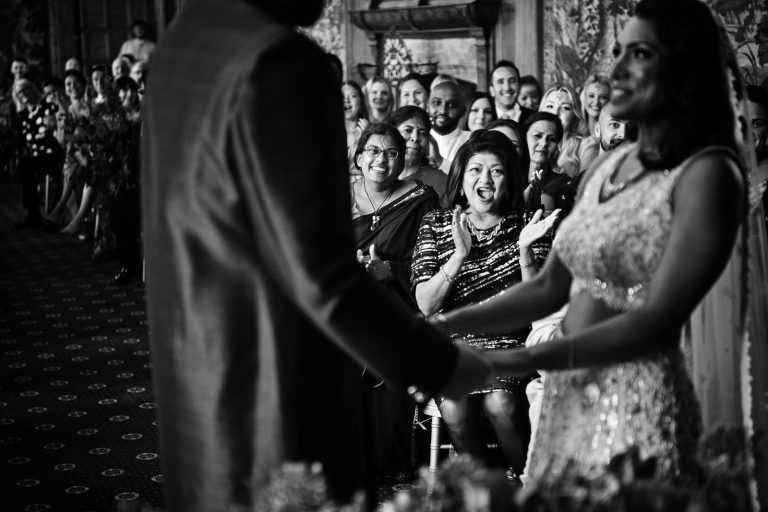 Wedding guests in the background clapping and cheering. Black and white image. Photographed at North Mymms Park, by Blooming Photography.)