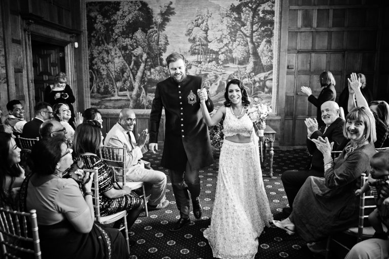 Bride and groom smiling as they walk down the wedding isle after their wedding ceremony, guests are smiling and cheering in the background. Black and white image. Photographed at North Mymms Park, by Blooming Photography.)