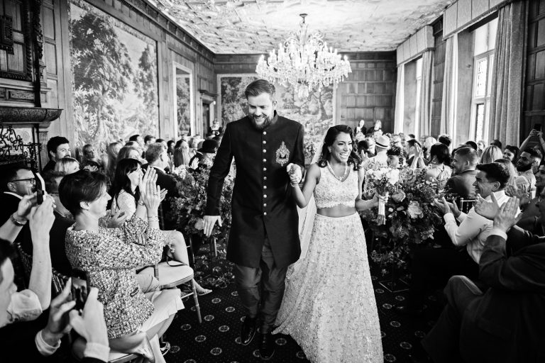 Bride and groom smiling as they walk down the wedding isle after their wedding ceremony, guests are smiling and cheering in the background. Black and white image. Photographed at North Mymms Park, by Blooming Photography.)