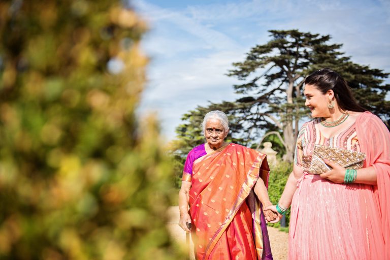Candid photograph of an elderly grandmother holding hands with a younger lady. Both dressed in stunning colourful sari's. Photographed at North Mymms Park, by Blooming Photography.