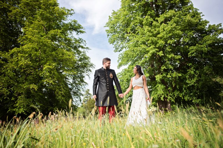 A bride and groom hold hands in the long grass with trees with fresh leaves behind them. Photographed at North Mymms Park, by Blooming Photography.