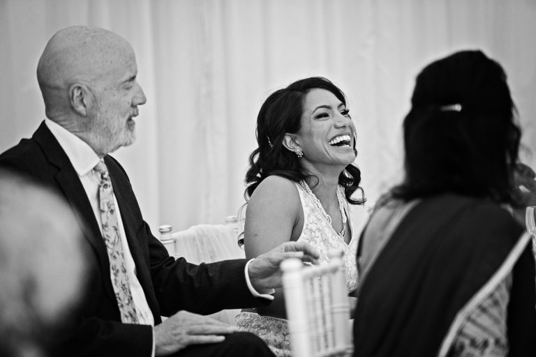 Bride laughs as she hears her groom making a funny wedding speech. Black and white photo. Photographed at North Mymms Park, by Blooming Photography.
