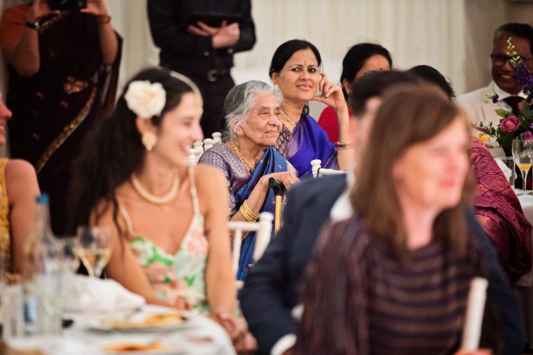 The grandmother of the bride listens to the grooms speech. She is smiling. Lots of other people surround her but all out of focus. Photographed at North Mymms Park, by Blooming Photography.