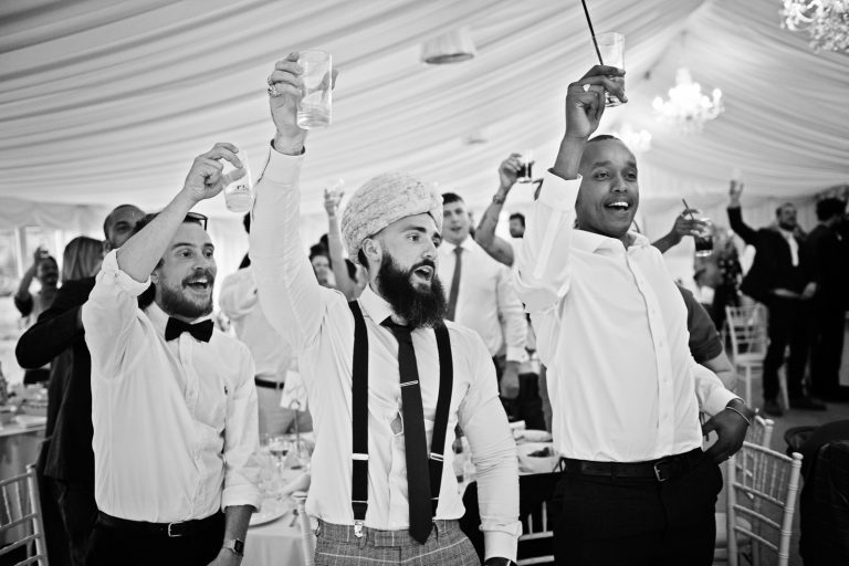 Guests stand and raise their glass, smiling and cheering at the end of the wedding speeches. Black and white photo. Photographed at North Mymms Park, by Blooming Photography.