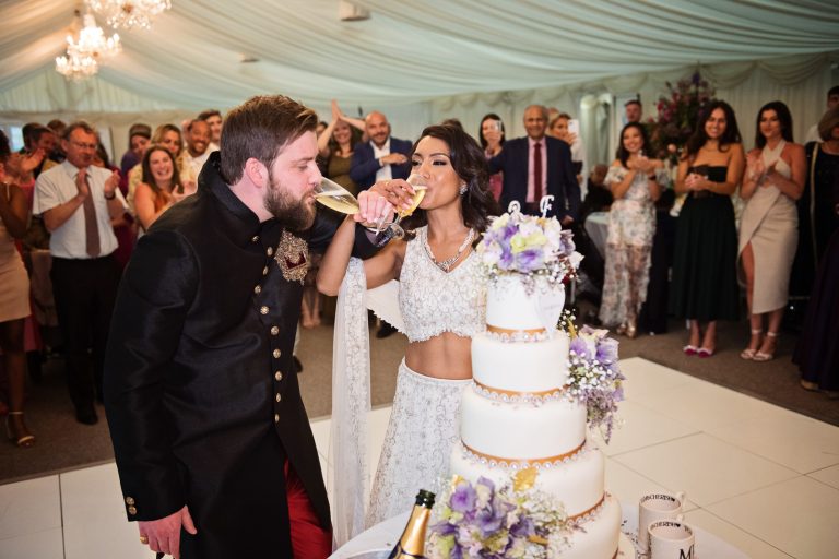 Bride and groom interlock their arms as they raise their glass of champagne and drink. Wedding guests behind them (out of focus) cheer too. They are standing next to the impressive wedding cake. Photographed at North Mymms Park, by Blooming Photography.