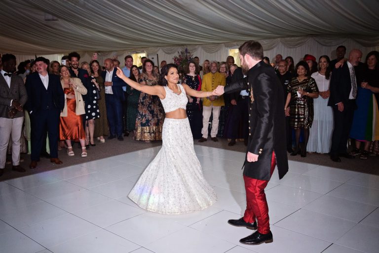 Bride and groom do their first dance. Bride and groom raise their glass and cheers each other as guests behind them (out of focus) cheer too. They are standing next to the impressive wedding cake. Black and white photo. Photographed at North Mymms Park, by Blooming Photography.