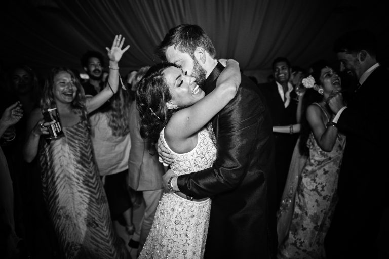 Bride and groom embrace as their guests cheer behind them. Photographed at North Mymms Park, by Blooming Photography.