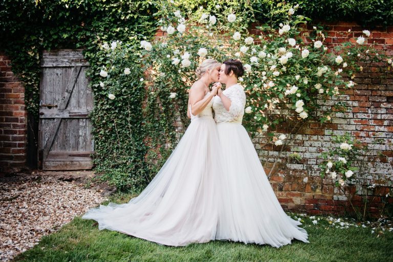 Bride and bride kiss, tummy's together and a white flowering rose against a red brick wall behind them.