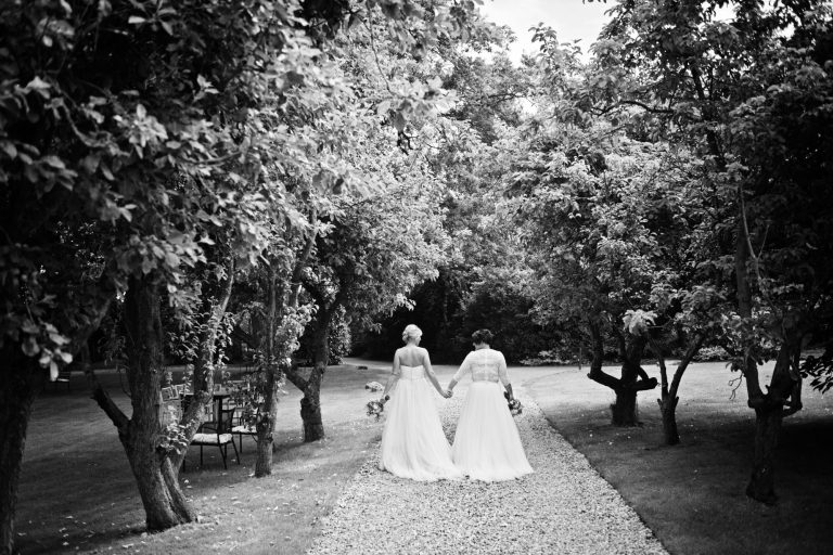 B&W photo of bride and bride holding hands and walking away from the camera down a tree lined driveway.