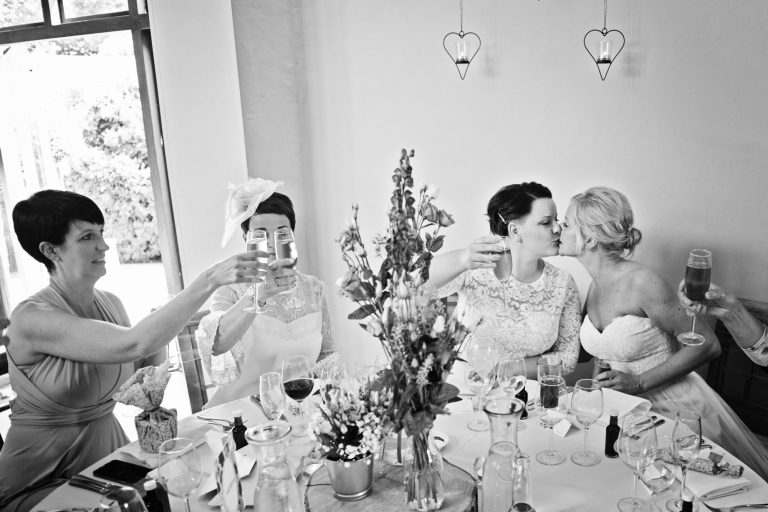 B&W candid photo of guests raising a glass whilst two brides kiss.