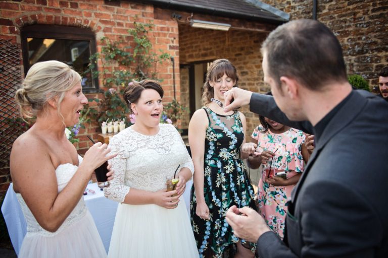 Colour photo of bride and bride watching a magic trick in awe.