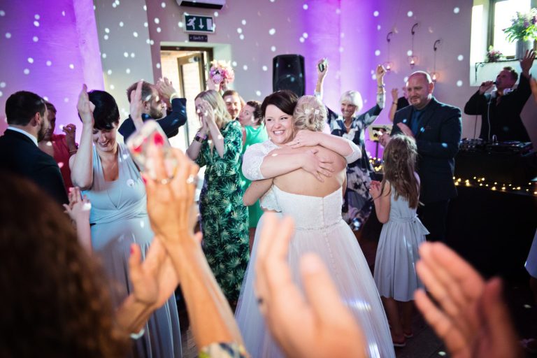 Bride and bride hug after their first dance with guests cheering and smiling surrounding them,